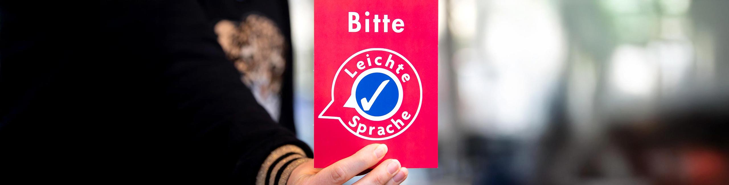 Jemand hält eine rote Karte in die Kamera. Auf der Karte steht 'Bitte Leichte Sprache'.