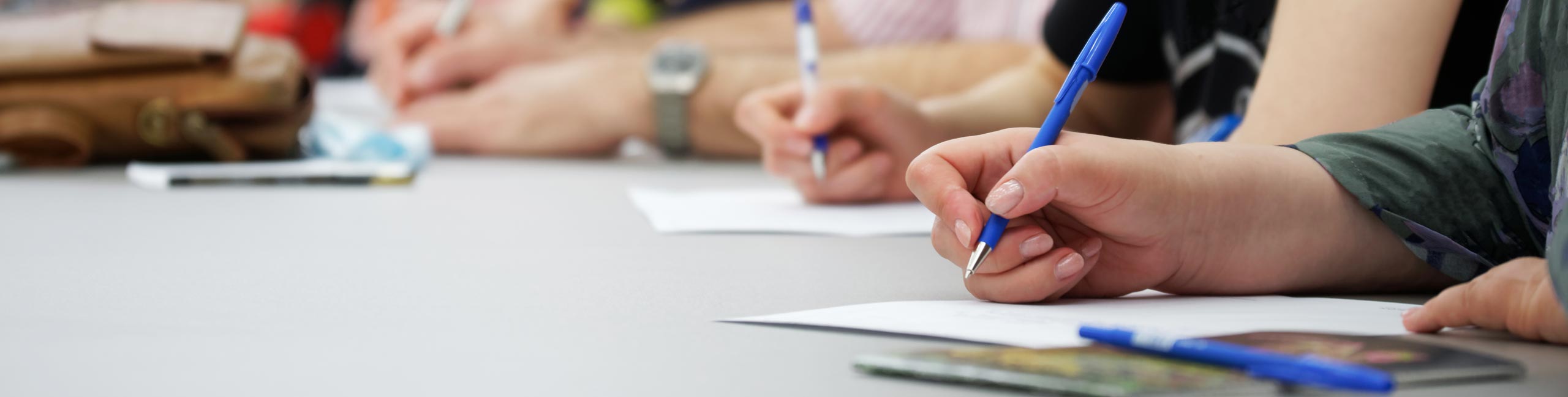 Eine Menschengruppe sitzt zusammen und schreibt etwas auf ein Papier. Man sieht nur die Papiere und die Hände, die einen Stift halten.